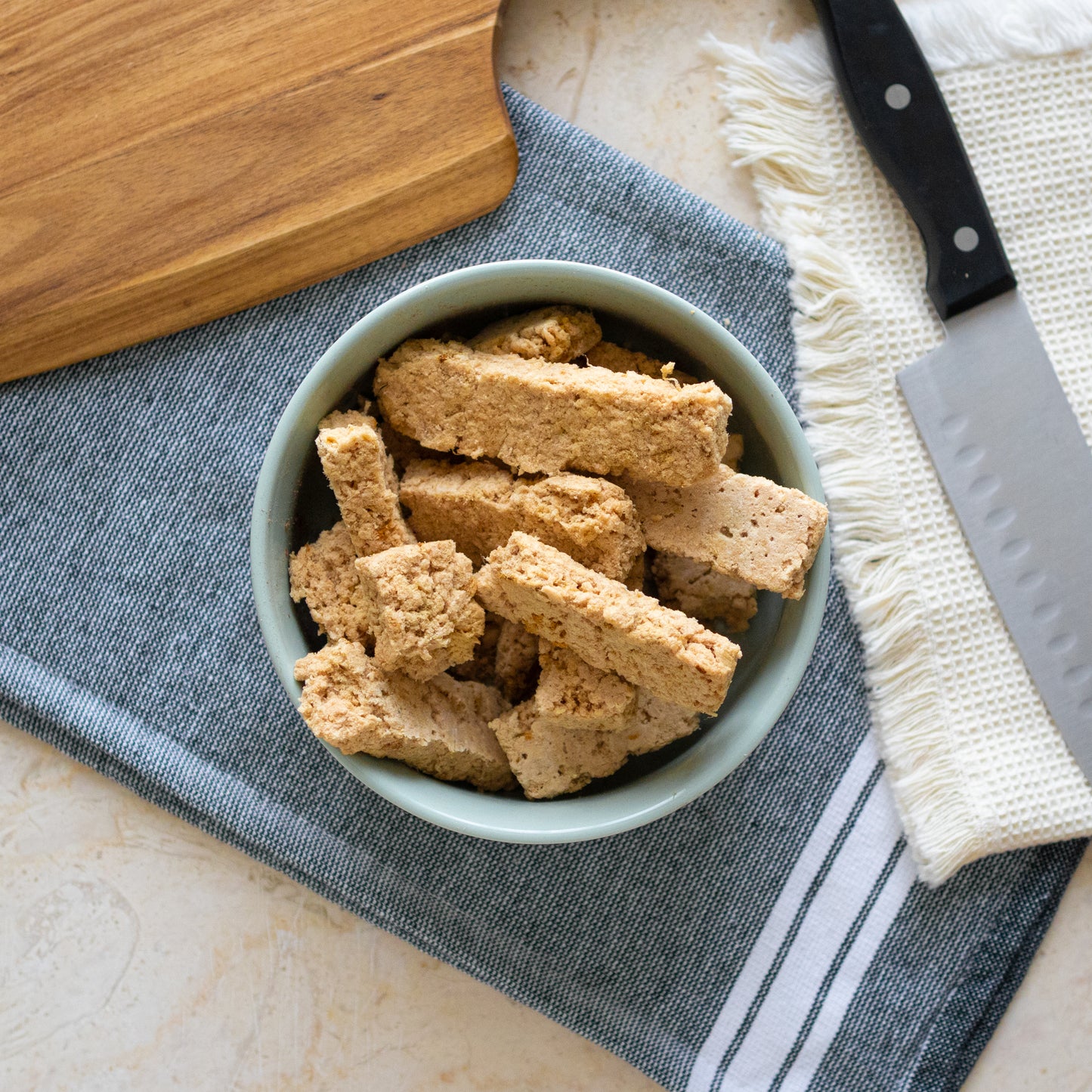 Bowl of Freeze Dried Raw Fish Sticks, a natural fish treat for dogs, displayed on a textured cloth with a wooden cutting board and knife in the background.